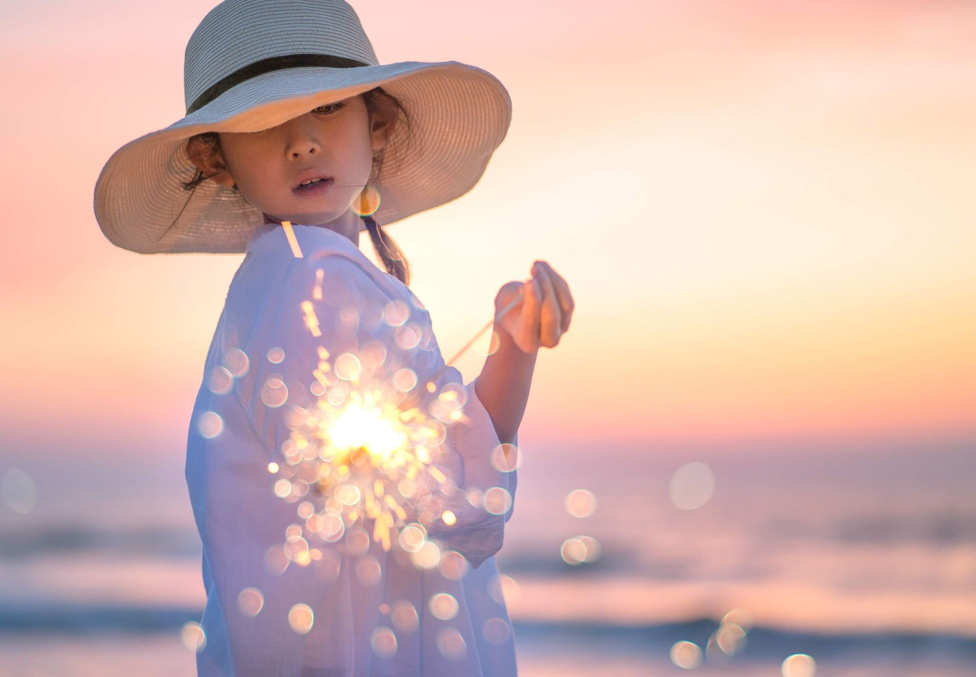 夕暮れの海辺で、麦わら帽子の子どもが手持ち花火を見つめる